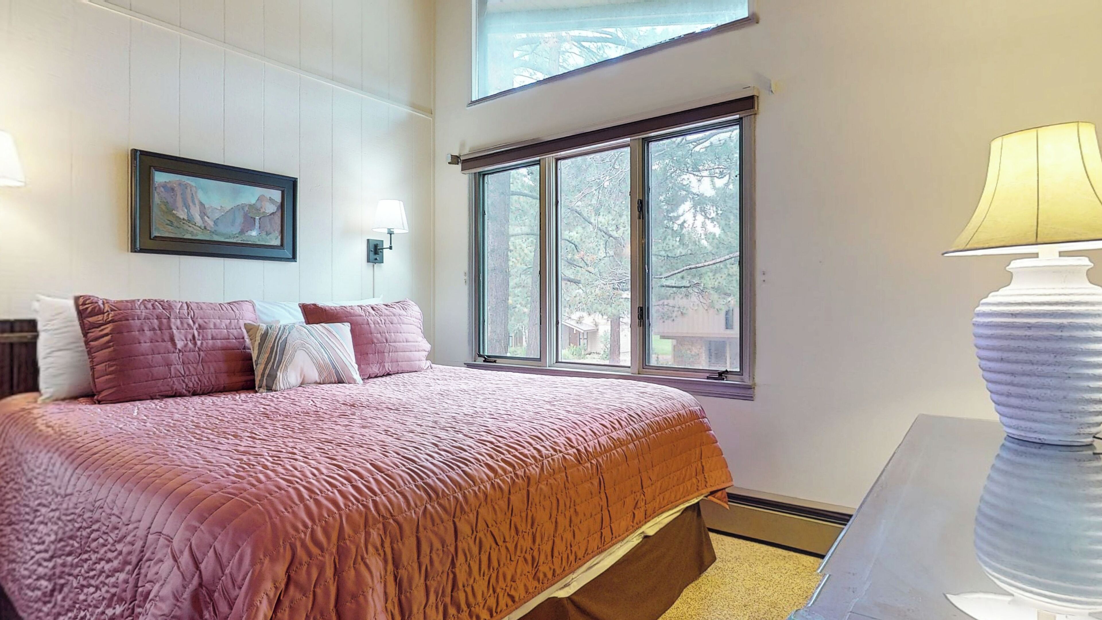 Quarto com uma cama grande, um quadro na parede, uma janela com vista para árvores e um abajur em uma mesa de cabeceira.