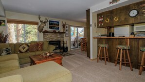 Een woonkamer met een open haard, een keukenhoek met krukjes, en een groot raam met uitzicht op bomen.