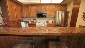 A kitchen with wooden cabinets, a stainless steel refrigerator, a microwave, and a stove. There are two plaid cushioned stools at the counter. - Mountainback #12, 2 Bdrm, 3 Ba, Loft - Pet OK (Mammoth Lakes)