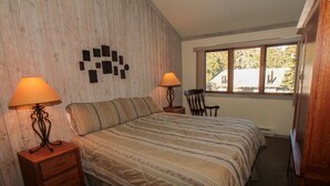 A bedroom with a bed, two bedside tables with lamps, a chair, a window with a view, and a wooden wall. - Mountainback #12, 2 Bdrm, 3 Ba, Loft - Pet OK (Mammoth Lakes)