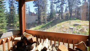 Blick aus einem Fenster mit Holzbalkon, auf einen bewaldeten Bereich mit Bäumen und einem Gebäude im Hintergrund.
