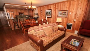 A cozy living room with a leather sofa, wooden dining table, and chairs. A coffee table with a magazine is in front of the sofa. The room has wooden walls and a kitchen in the background. - Mountainback #49, Den (Mammoth Lakes)