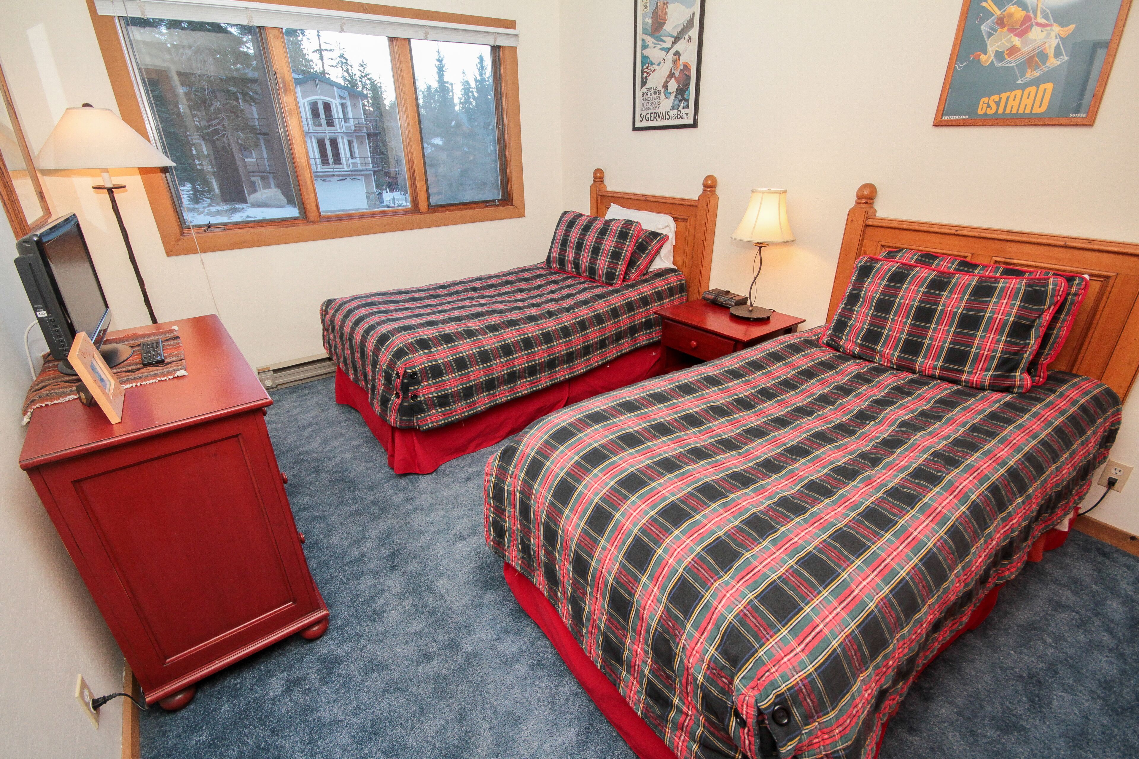 A bedroom with two beds, a desk, a chair, and a window with a view of a building and trees.