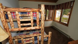 Uma cama de madeira rústica com roupa de cama xadrez, uma janela com cortinas e uma luminária.