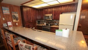 A kitchen with wooden cabinets, a granite countertop, a microwave, a refrigerator, and bar stools. - Mountainback #105, Loft, Den (Mammoth Lakes)