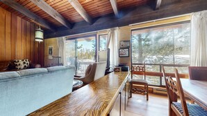 Une pièce chaleureuse avec un plafond en bois, un canapé, une table à manger avec des chaises et de grandes fenêtres donnant sur des arbres et un paysage enneigé.