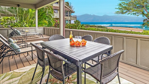 Un espace extérieur abrité, aménagé pour les repas, avec une table pour deux, entouré de chaises en osier, et offrant une vue sur une plage avec des montagnes à l’horizon.