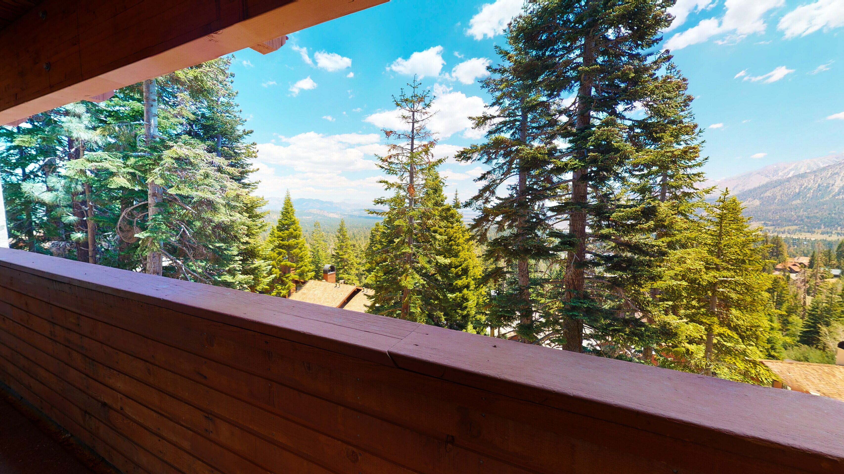 A wooden balcony with a view of tall evergreen trees and distant mountains.