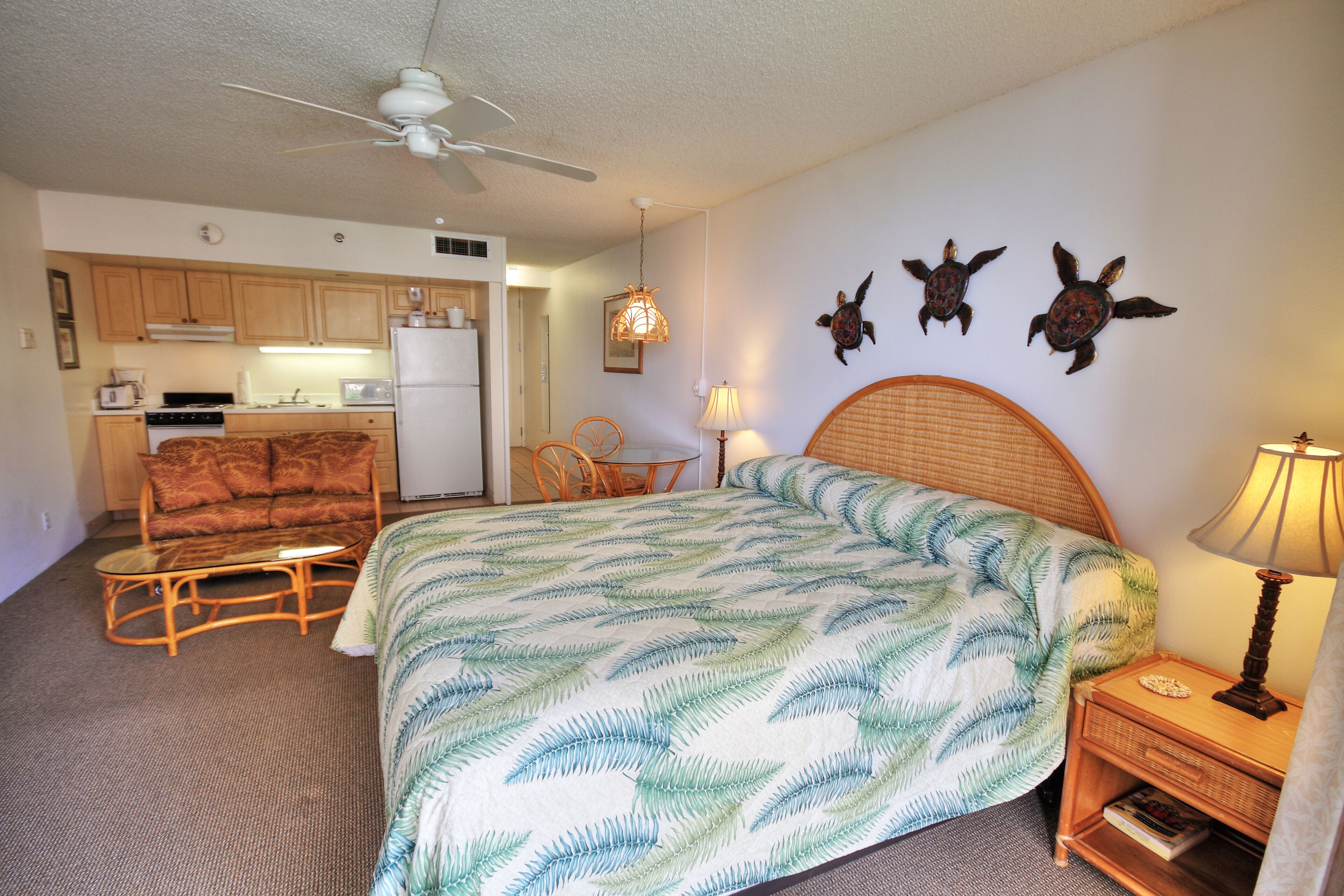 Une chambre d’hôtel de taille modeste, comprenant un lit, une chaise, une petite table, une lampe et, au fond, une kitchenette.