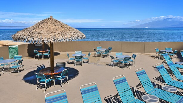 Eine Strandlounge mit Liegestühlen und Tischen, einem Reetdachschirm und Blick auf den Ozean.