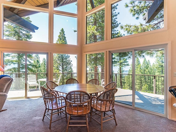 A spacious room with a large round table and six chairs, large windows, and a balcony with a view of trees.