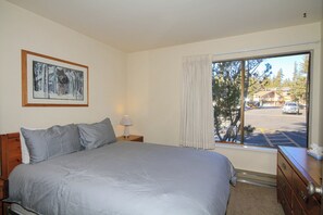 Une chambre à coucher comprenant un lit, une table de chevet, une lampe, une commode, une fenêtre donnant sur un parking et des arbres, ainsi qu’un tableau encadré accroché au mur.