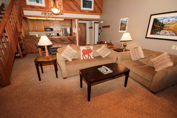 A cozy living room with a sofa, coffee table, and lamps. A kitchen is visible in the background. - Mountainback #53, Loft (Mammoth Lakes)