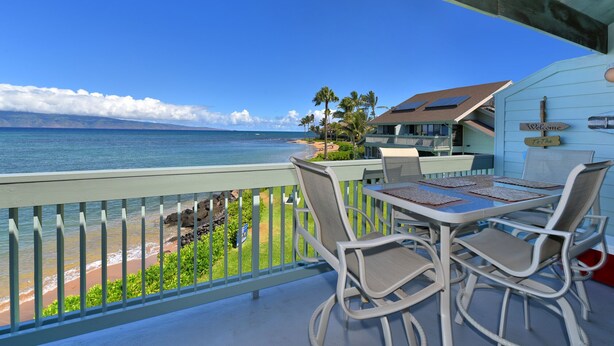A balcony with a table and chairs overlooking a beach with palm trees and a house in the background.
