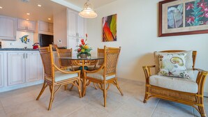 A dining area with a round table and four wicker chairs, a kitchen with white cabinets, and a living area with a wicker sofa and a framed picture on the wall. - Vitamin Sea available on Maui Come Play at Kahana Reef 402   (Lahaina)