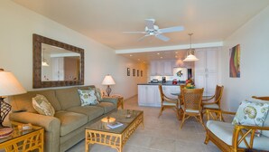 A living room with a sofa, coffee table, and dining area with chairs. There is a kitchen in the background with cabinets and a ceiling fan. - Vitamin Sea available on Maui Come Play at Kahana Reef 402   (Lahaina)
