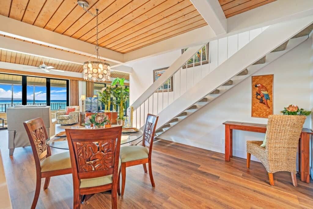 A spacious living area with a wooden floor, a dining table with chairs, a staircase, and a view of the ocean.