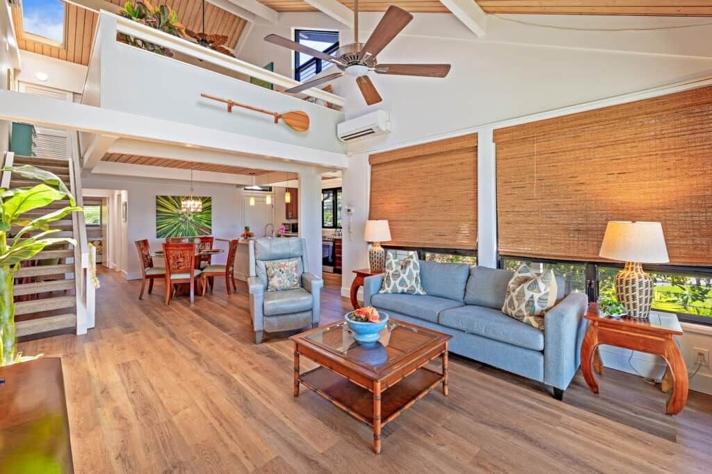 Un vaste espace de vie comprenant un canapé bleu, une table basse en bois et une table à manger avec des chaises. On peut également apercevoir un ventilateur de plafond et une fenêtre offrant une vue agréable.