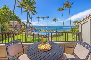 A balcony with a table set for two, overlooking a pool and palm trees, with a view of the ocean. - Kahana Village #14 Beach Front Luxury (Lahaina)