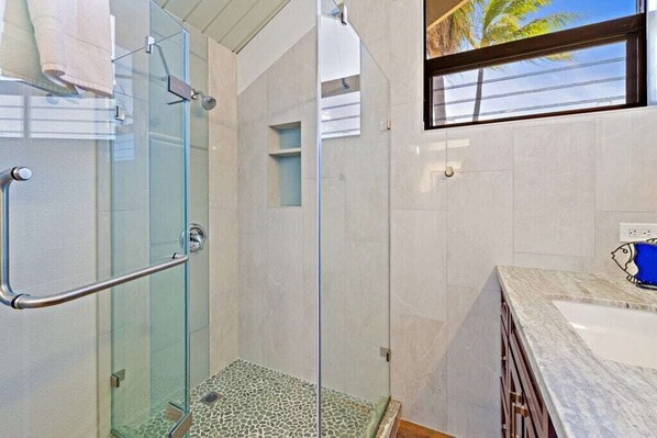 A modern bathroom with a glass shower enclosure, a marble countertop, and a view of palm trees through a window. - Kahana Village #14 Beach Front Luxury (Lahaina)