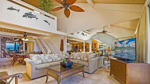 A spacious living area with a large sofa, a coffee table, and a television. There is a staircase leading to an upper level, and the room has a ceiling fan. - Kahana Village #21 Beach Front Luxury (Lahaina)