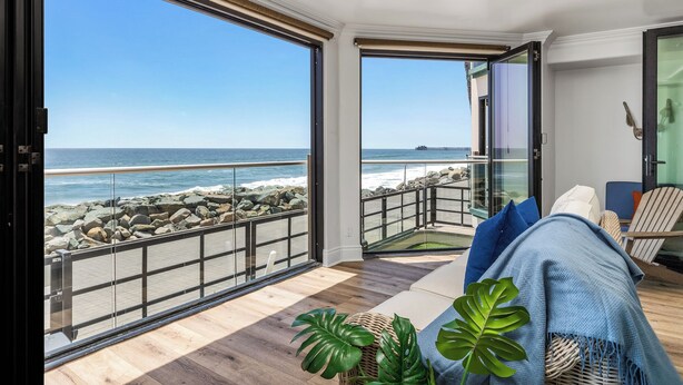 Une pièce dotée d’une grande porte vitrée donnant sur un balcon avec vue sur l’océan, d’un plancher en bois et d’un coin salon avec des coussins.