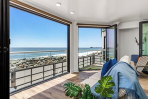 Une pièce dotée d’une grande porte vitrée donnant sur un balcon avec vue sur l’océan, d’un plancher en bois et d’un coin salon avec des coussins.