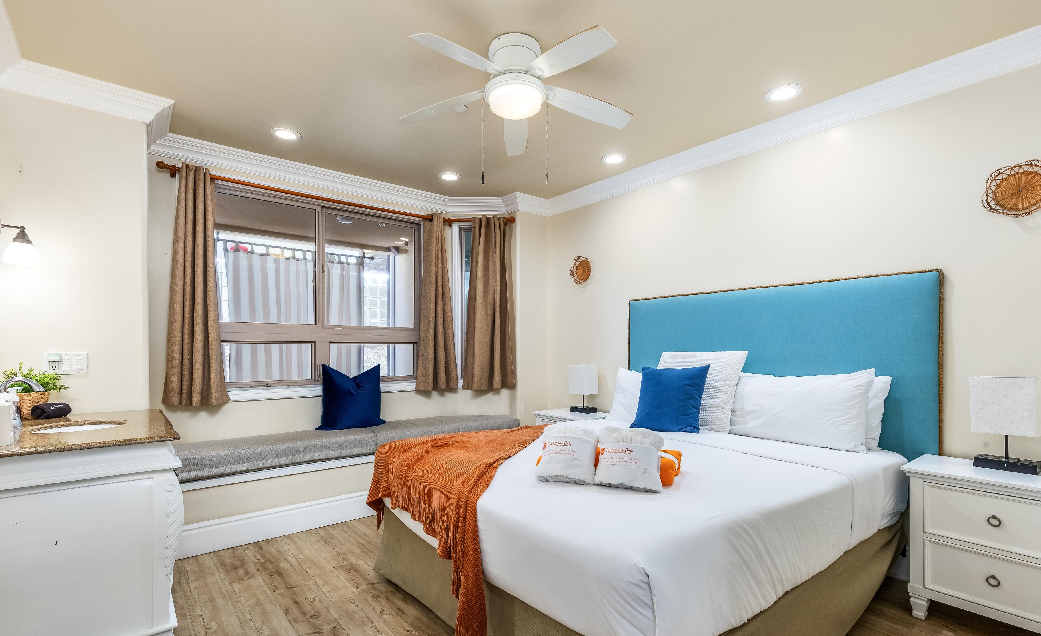 Habitación de hotel con cama, mesita de noche, ventana con cortinas, ventilador de techo y un reloj de pared.