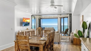 Un coin repas avec une table et des chaises en bois, un ventilateur de plafond et une vue sur l’océan grâce à de grandes fenêtres.