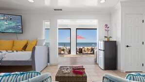 A living room with a sofa, a coffee table with a puzzle, a TV displaying a beach scene, and a door leading to a balcony with beach chairs and a red umbrella. - Single Family Gorgeous Beachfront Mansion on the Sand P1733-0 (Oceanside)