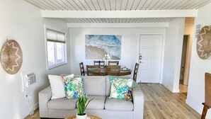 Een woonkamer met een grijze bank, een rond rieten bijzettafeltje, en een eethoek met een houten tafel en stoelen.