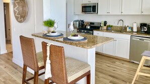 Een moderne keuken met witte kastjes, een magnetron, een fornuis en een vaatwasser. Er staan twee rieten stoelen met kussens en een klein eiland met een schaal fruit en een bloevas.