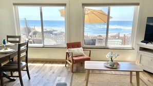 A room with a wooden floor, a dining table with chairs, a sofa, a coffee table, and a TV. Large windows with a view of the ocean and beach furniture are visible. - Oceanfront Getaway with Private Sandy Beach - 1615-1 (Oceanside)