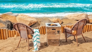 A beachside seating area with two wicker chairs, a small wooden table with a fire pit, and a view of the ocean. - Oceanfront Getaway with Private Sandy Beach - 1615-1 (Oceanside)