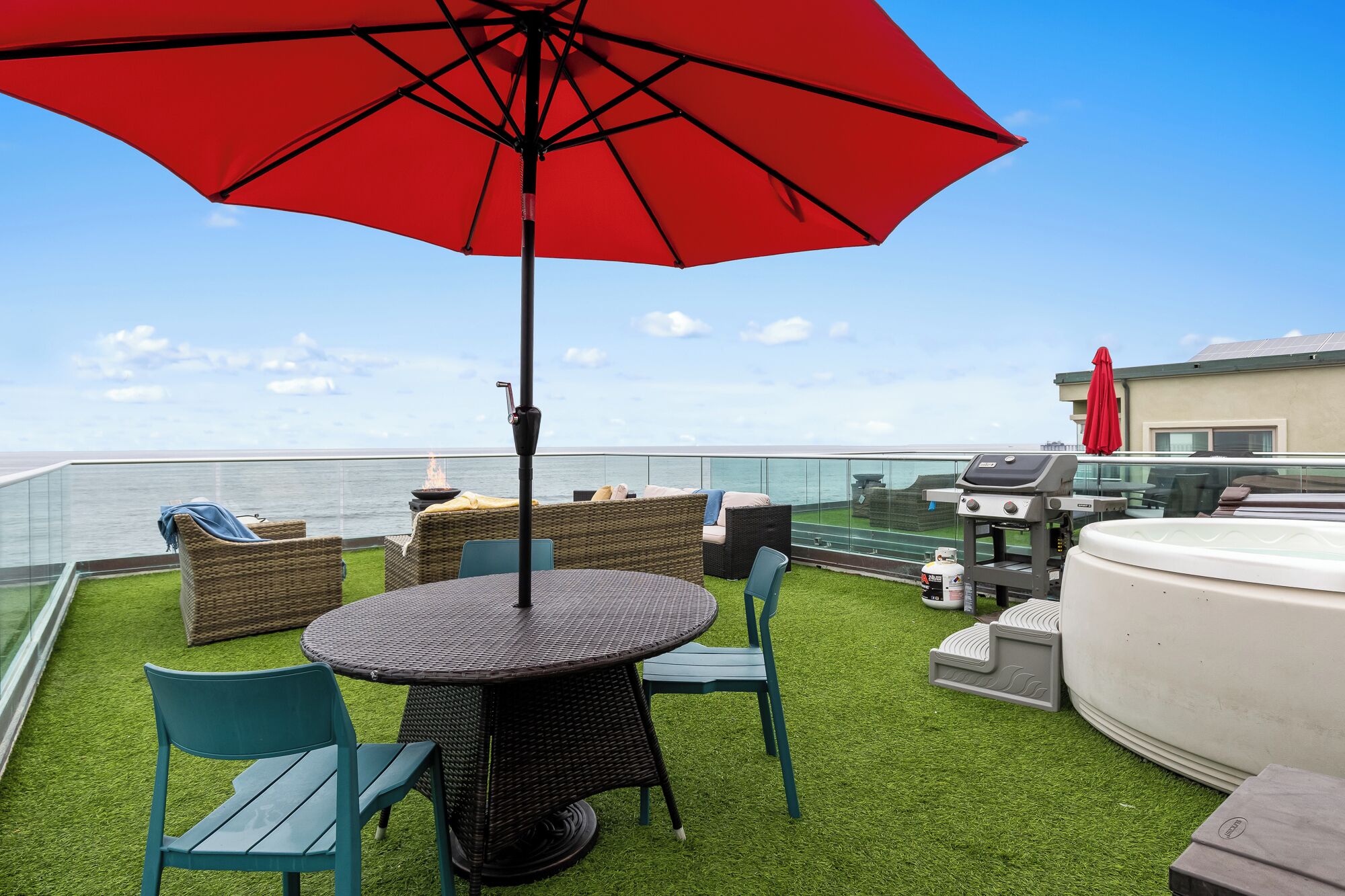 Una terraza en la azotea con una mesa redonda, sillas, una sombrilla roja, una parrilla y vistas al océano.