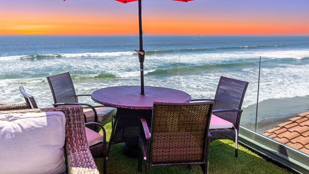Une terrasse en bord de mer avec une table ronde et des chaises, un parasol rouge et une vue sur l’océan au coucher du soleil.