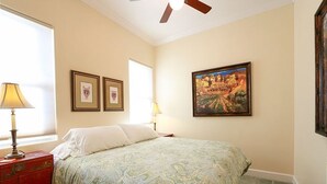 Un dormitorio con cama, mesita de noche, lámpara, ventilador de techo y un cuadro en la pared.