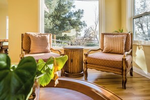 A room with two wooden chairs with patterned cushions, a round wooden table, and a view of trees through a large window.