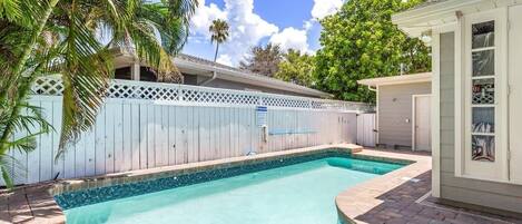 A swimming pool with a wooden deck, a white fence, and a small building with glass doors.