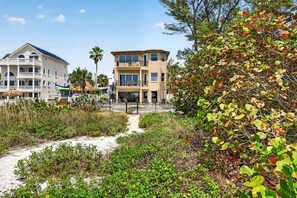 A coastal residential area with two multi-story houses, a sandy path, and dense vegetation.