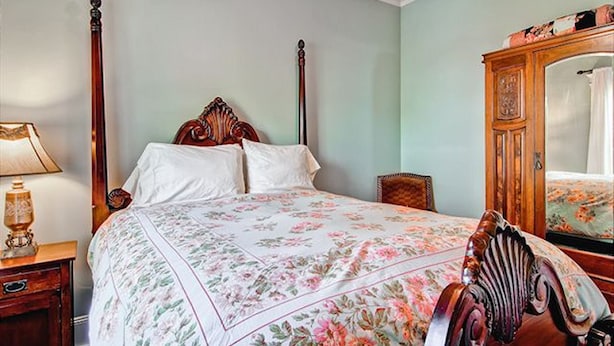 A bedroom with a four-poster bed, a wooden nightstand with a lamp, a chair, and a mirror on the wall.