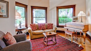 A living room with a sofa, a coffee table, a patterned rug, a piano, and large windows with a view of greenery.