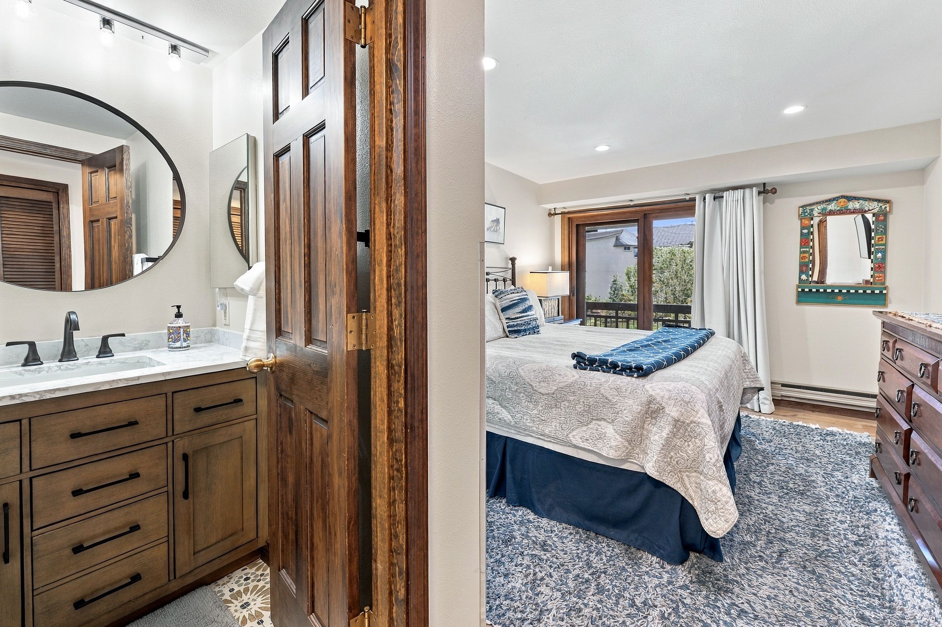 Un dormitorio con cama, cómoda, ventana con cortinas y baño con lavamanos y espejo.