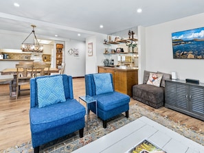 A modern living room with two blue armchairs, a grey sofa, and a kitchen area with wooden cabinets and a hanging light fixture. - Walk to Riverfront Gondola (Avon)