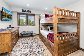 A bunk bed with a wooden frame, a bedside table with drawers, a bench with a patterned cushion, a window with blinds, and a wall-mounted picture.