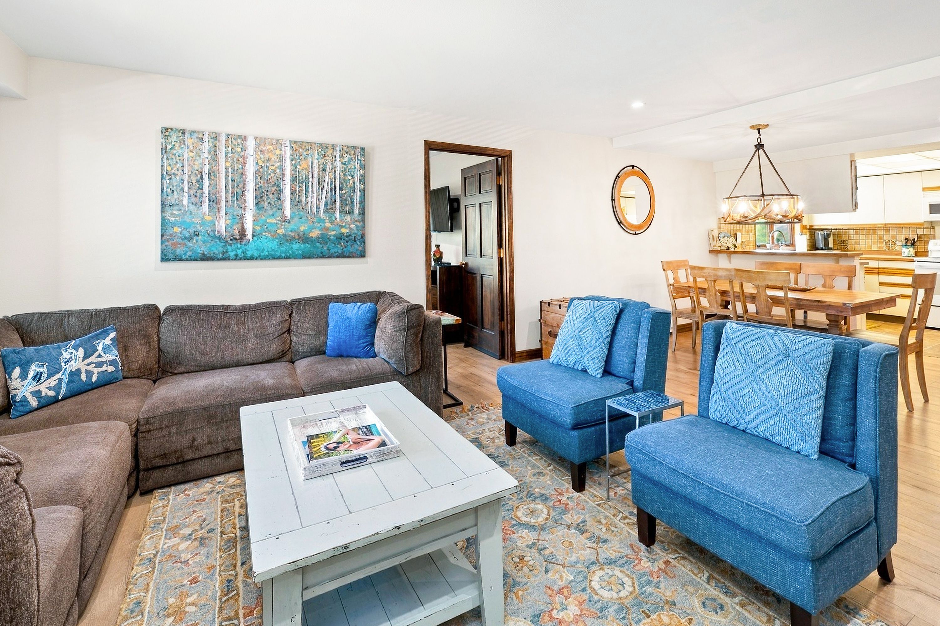 A living room with a sofa, a coffee table, and a blue armchair. There is a painting on the wall and a dining area with a table and chairs.