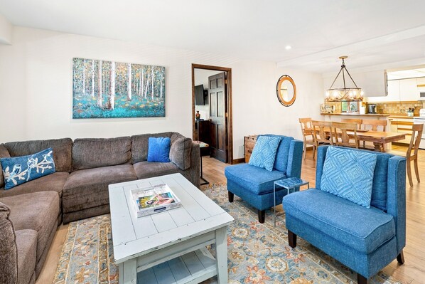 A living room with a sofa, a coffee table, and a blue armchair. There is a painting on the wall and a dining area with a table and chairs.