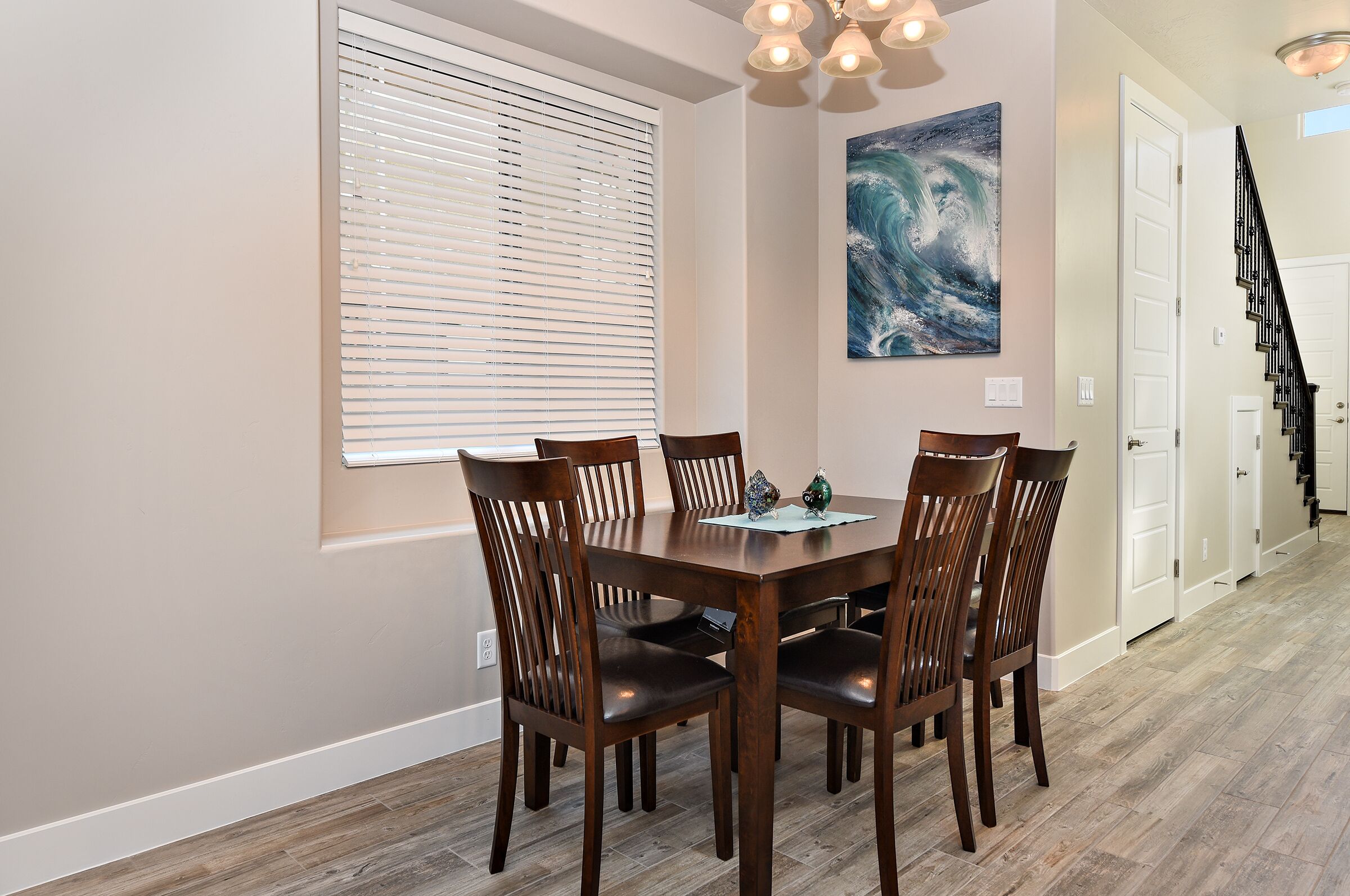 Un área de comedor con una mesa y sillas de madera, una ventana con persianas, un cuadro en la pared y una escalera al fondo.