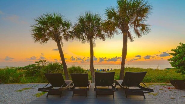 Una terraza con sillones de descanso frente a una vista al atardecer, palmeras y una playa en el fondo.