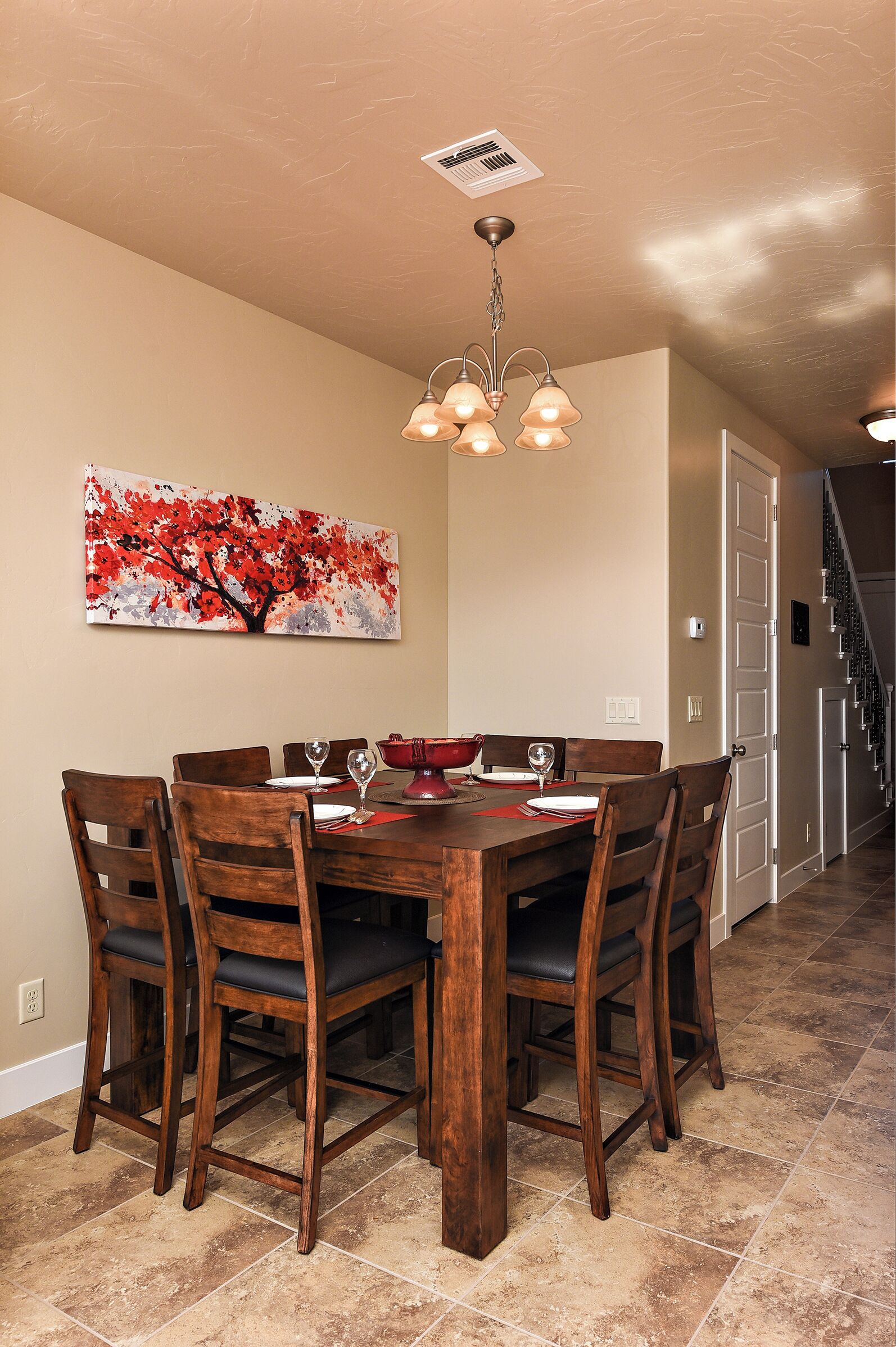 Un coin repas avec une table en bois, des chaises et un lustre. Un tableau orne le mur, et un escalier se trouve au fond de la pièce.